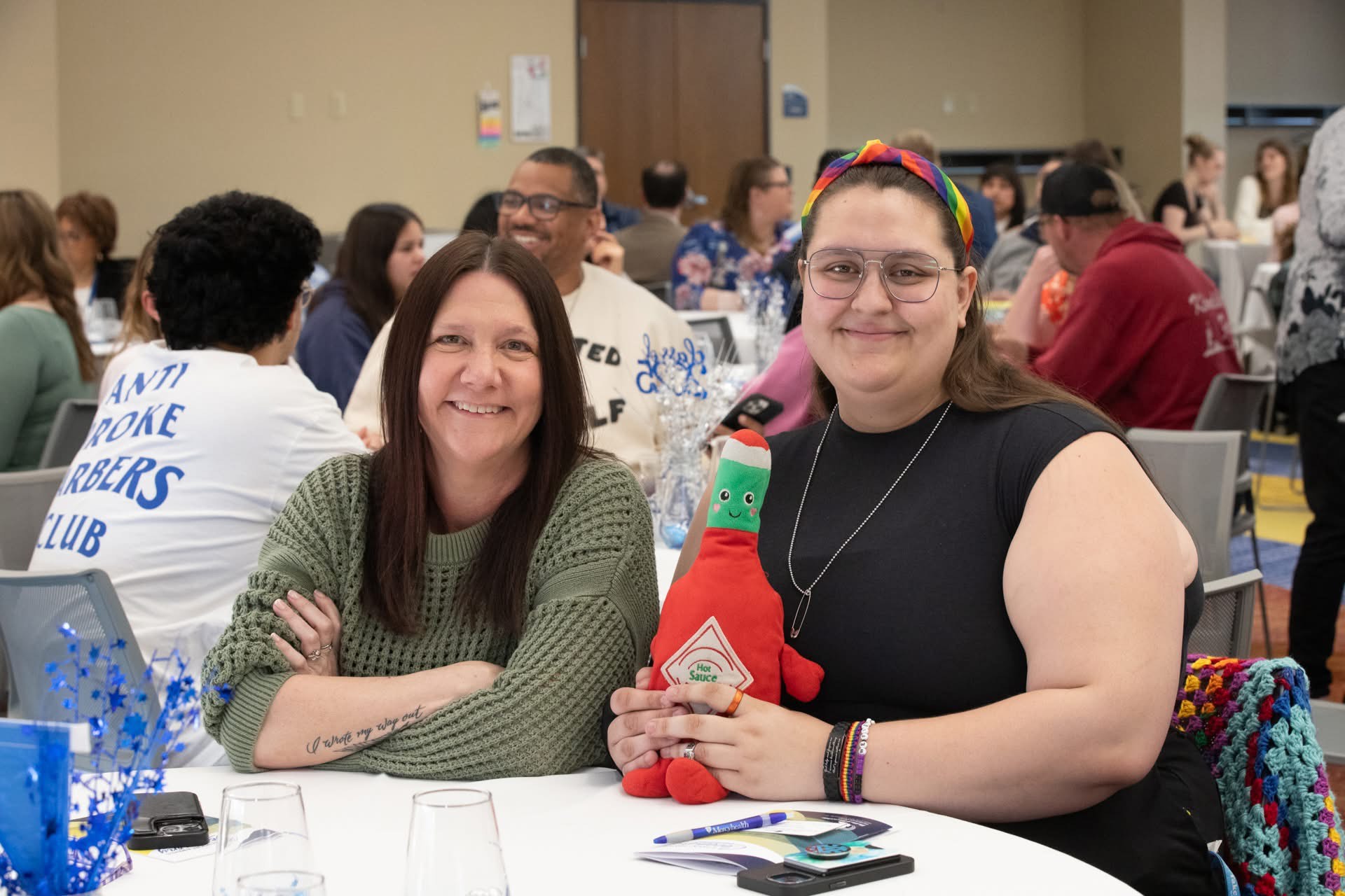 A photo of 2 women sitting at a table with a white tablecloth. The woman on the left is in a green knit sweater and smilling. The woman on the right is in a black sleeveless top and is holding a hot sauce plushie in a sitting position on the table. 